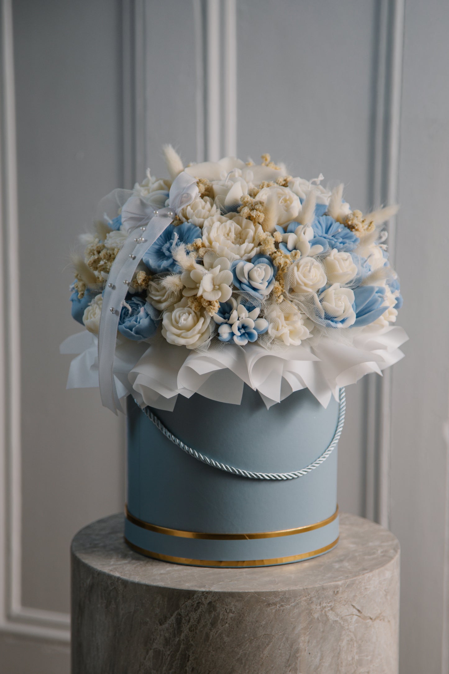 Decorative flower arrangement in a blue container on a gray pedestal against a gray paneled wall.